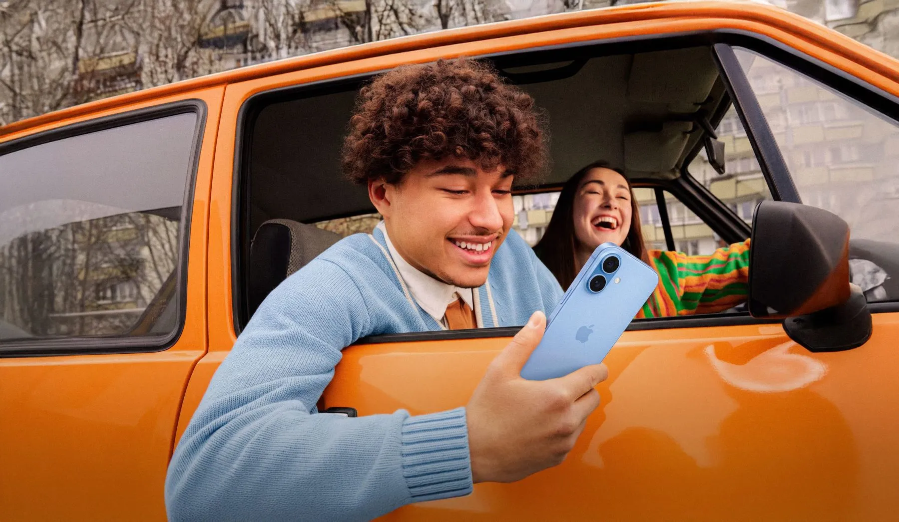Zwei Freunde lachen in einem orangen Auto, einer schaut aufs Handy