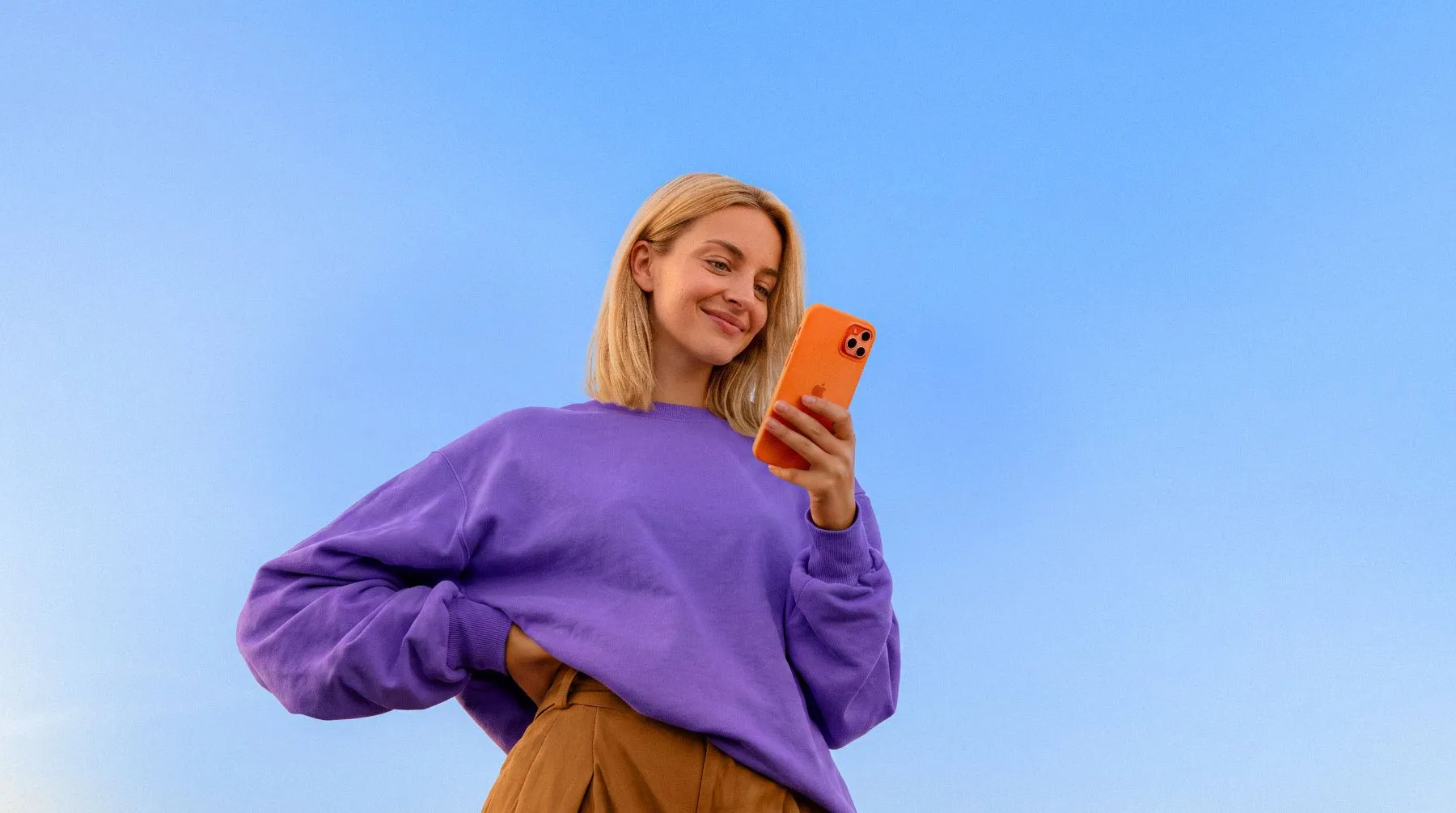 Lächelnde Frau im lila Pullover blickt vor blauem Himmel auf ihr oranges Handy